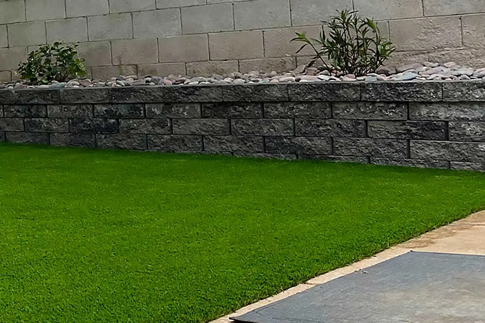 A neatly designed Las Vegas landscaping project features a lush artificial grass lawn bordered by a stone retaining wall with drought tolerant plants and decorative pebbles, creating a low maintenance, visually appealing outdoor living space.| Sun Valley Landscape Las Vegas