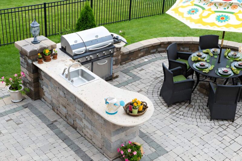 Outdoor kitchen area with a built-in grill, sink, and round dining table set for six under a colorful umbrella in a paved yard.