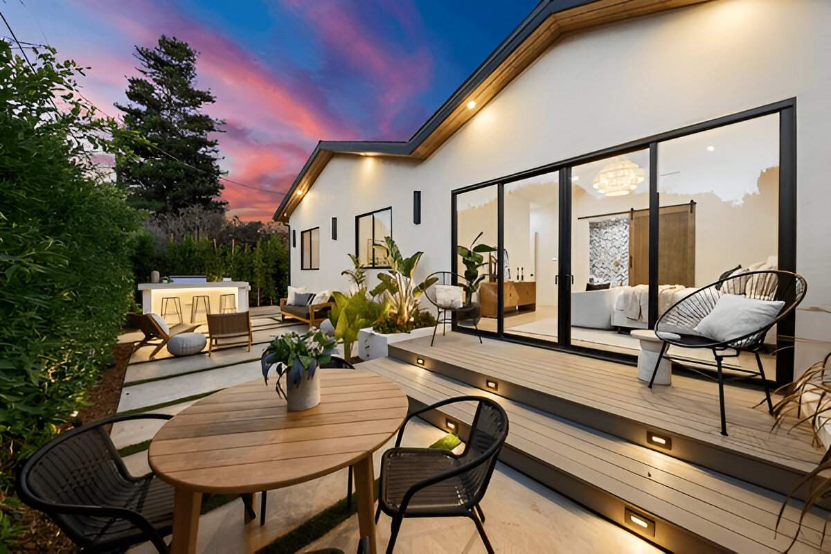 Modern outdoor patio with table, chairs, greenery, and cozy lighting at sunset, adjacent to a house with large glass doors.