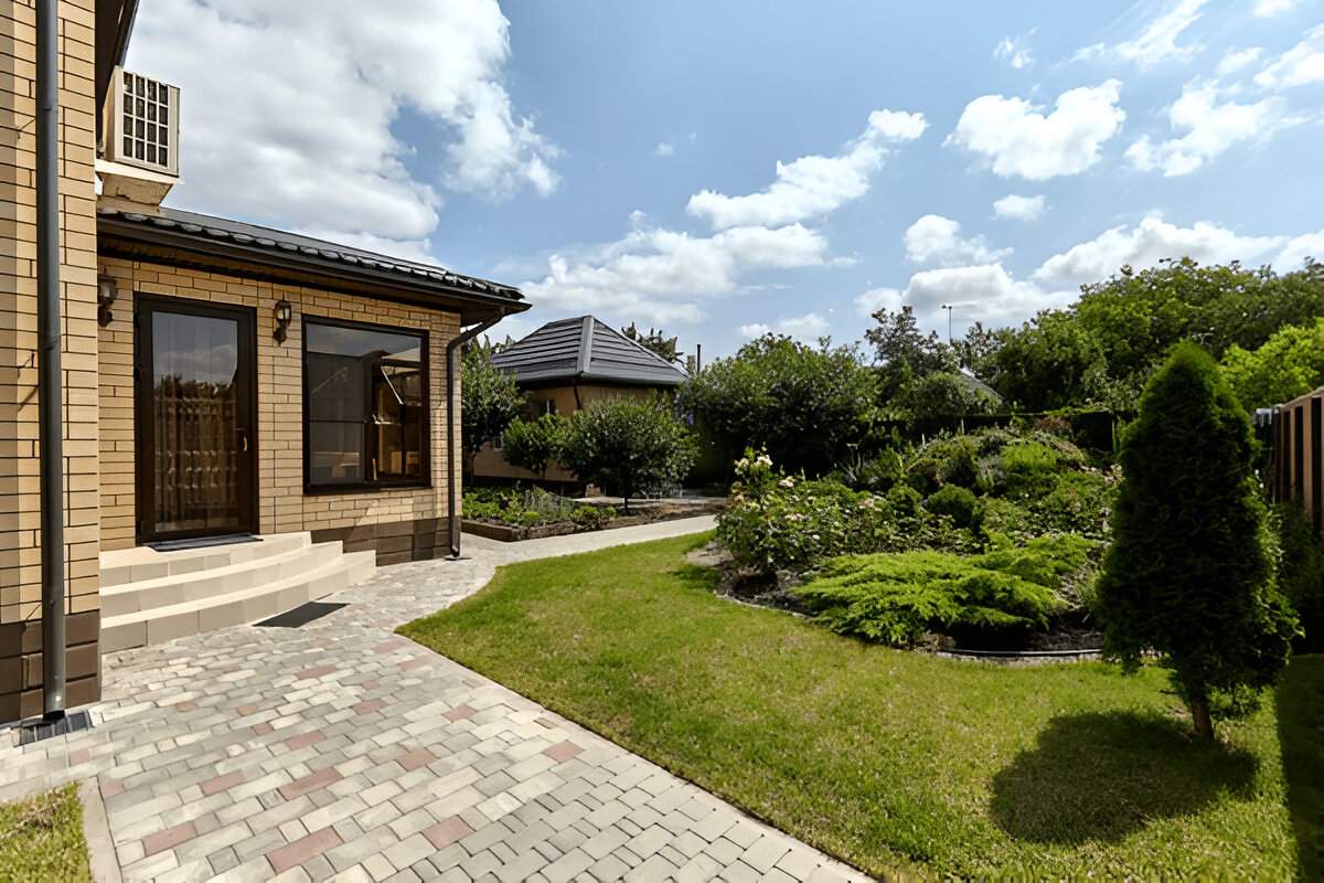 Paved walkway and manicured garden with lush greenery by a tan brick house under a blue sky with scattered clouds.
