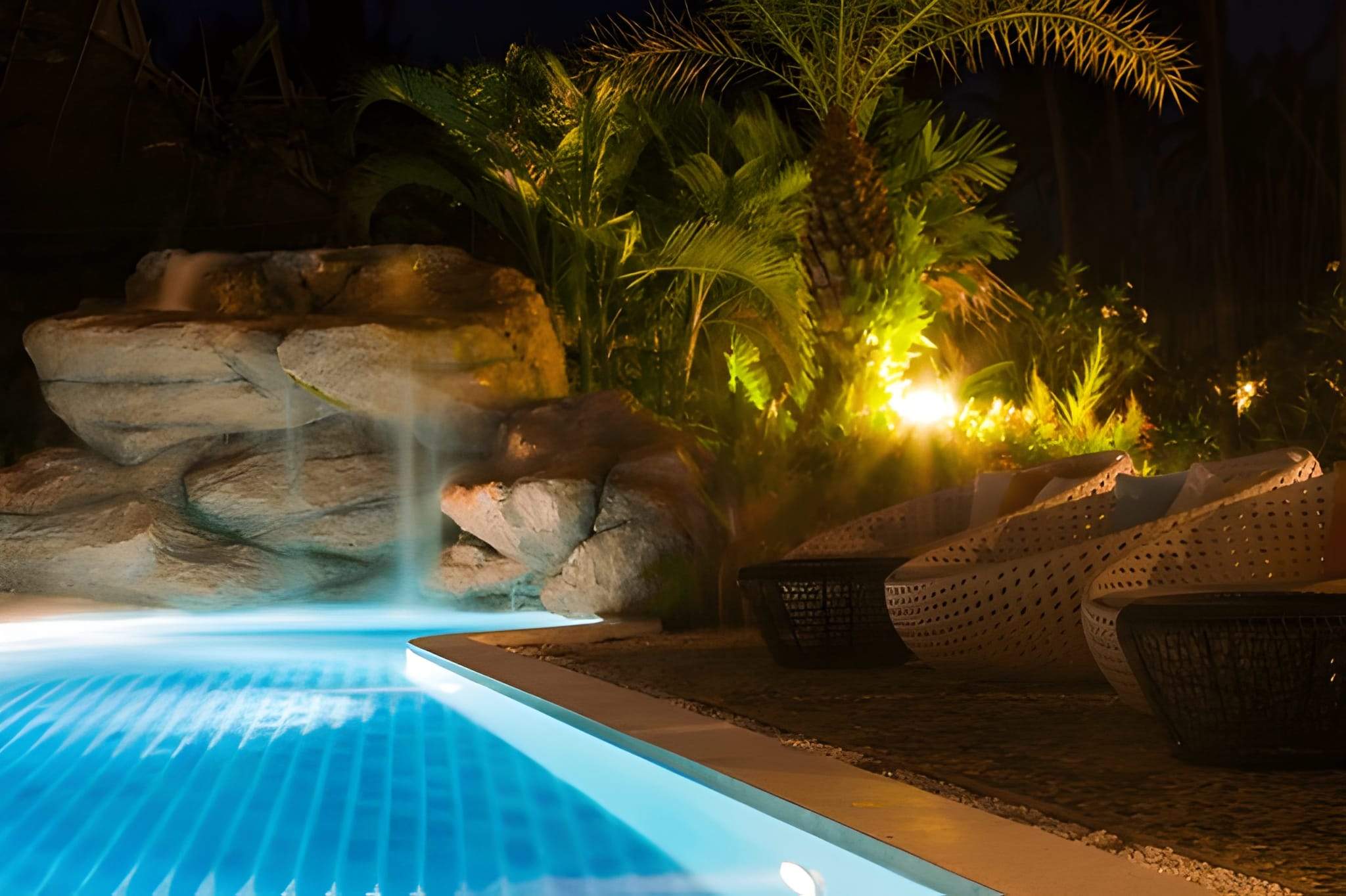 Poolside landscape lighting with palm trees and waterfall feature glowing under warm lights.